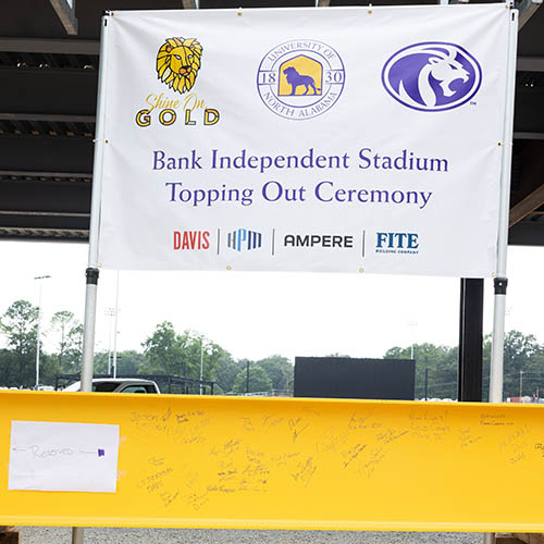 sign with una shine on gold logo, una logo, athletics logo, spongers, and text saying 'bank independent stadium topping out ceremony'