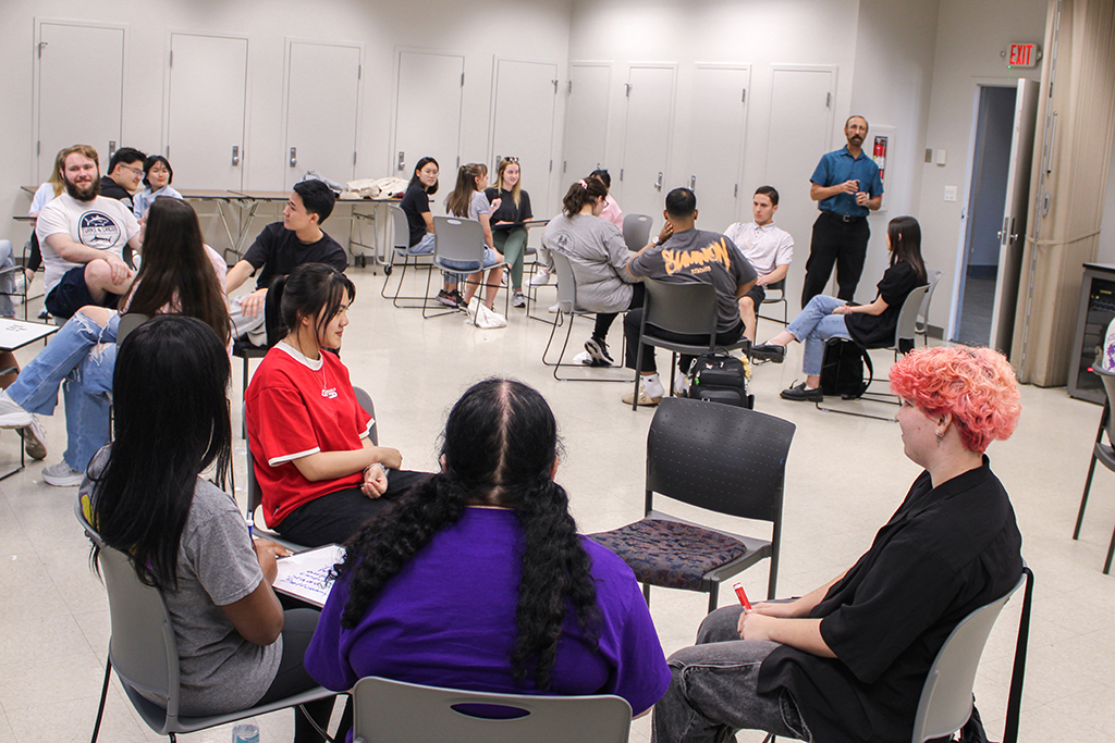 Full room of students talking in seated groups at Converation Partners meeting.